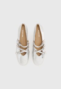 Scarpe in ecopelle argentata con cinturini incrociati e un design a taglio sulla parte anteriore. Suola piatta e interno foderato in materiale marrone chiaro.