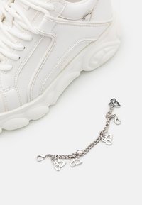 Weiße Ledersneaker mit strukturierten Akzenten, abgerundeter Sohle und einem dekorativen silbernen Schmetterlingsanhänger-Armband an der Seite.