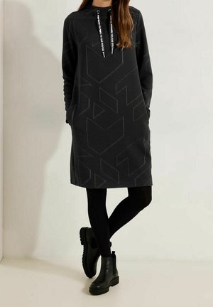Femme portant une robe à capuche noire à motif géométrique, leggings noirs et bottines noires épaisses, debout sur un fond clair uni.