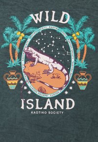 Mørkegrøn T-shirt med et grafisk design med palmer, en firben på en sten og dekorativ keramik. Teksten lyder "WILD ISLAND" og "KAOTIKO SOCIETY."