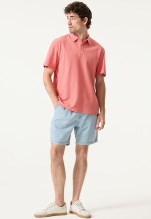 Korallenrotes Polohemd mit einer strukturierten Webart, Dreiknopf-Leiste, kombiniert mit hellblauen Jeansshorts und beigen Sneakers.