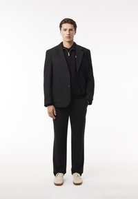 Chaqueta de traje negra con dos botones, polo negro con cremallera debajo y pantalones negros, combinados con zapatillas crema. Fondo liso.