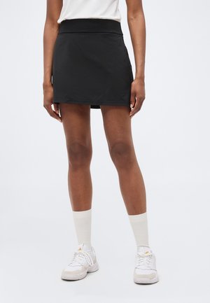 Femeie purtând o fustă sport neagră, șosete albe, adidași albi și un top alb, stând pe un fundal simplu și deschis la culoare.