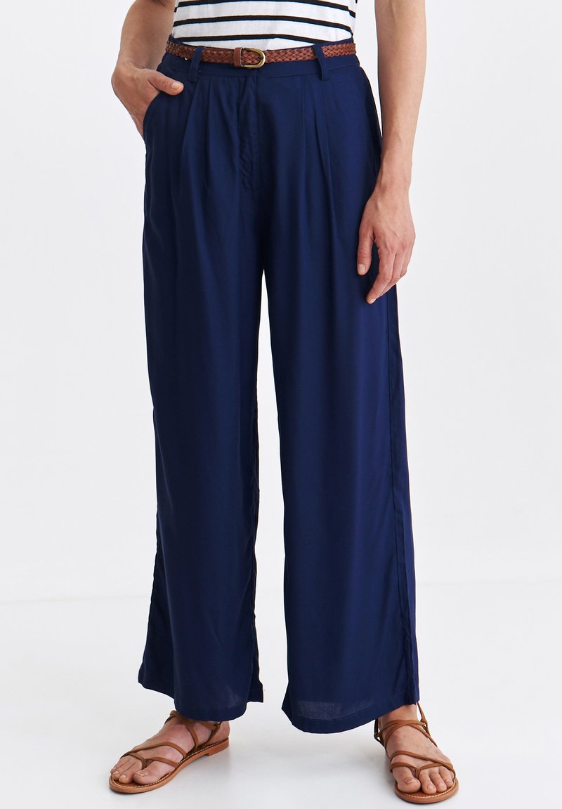 Granatowe spodnie z szerokimi nogawkami wykonane z lekkiej tkaniny, z plisowanym przodem i brązowym plecionym paskiem. Dopełnione brązowymi sandałami na paski.