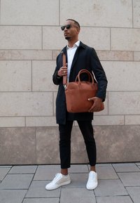 Braune Leder-Laptoptasche mit zwei Griffen und einem Umhängegurt, glatte Oberfläche, rechteckige Form. Getragen von einer Person in einem dunkelfarbenen Mantel und einem weißen Hemd.