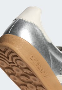 Närbild av en silver- och vit Adidas-sneakerhäll med en gummisula som har cirkulära mönster för grepp och Adidas-logotyp.