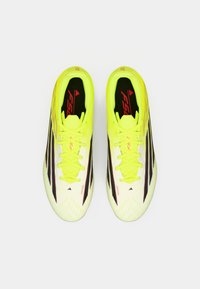 Φωτεινά κίτρινα ποδοσφαιρικά παπούτσια με μαύρες λεπτομέρειες και υφασμάτινο μοτίβο. Διαθέτουν κορδόνια και σήμα "FS 1" στη γλώσσα και στο πλάι.