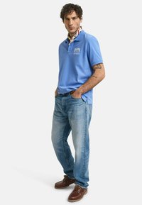 Ljusblå polotröja med broderad logotyp, tillsammans med blekta blå jeans och bruna snörskor. Subtila texturerade detaljer på polotröjan.