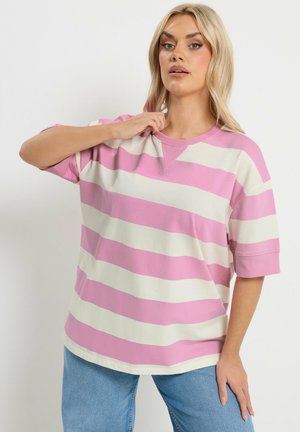 Kvinde med blondt hår iført en oversized pink og hvidstribet kortærmet skjorte og blå jeans, som justerer kraven med den ene hånd.