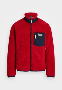Giacca in pile rossa con colletto alto, fronte con zip, dettagli blu navy e una tasca con chiusura a zip. Tessuto testurizzato con un aspetto morbido.
