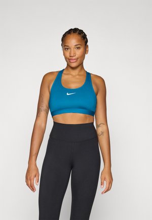 Een vrouw met ingevlochten haar glimlacht, gekleed in een teal sportbeha en zwarte leggings, met een zelfverzekerde atletische houding.
