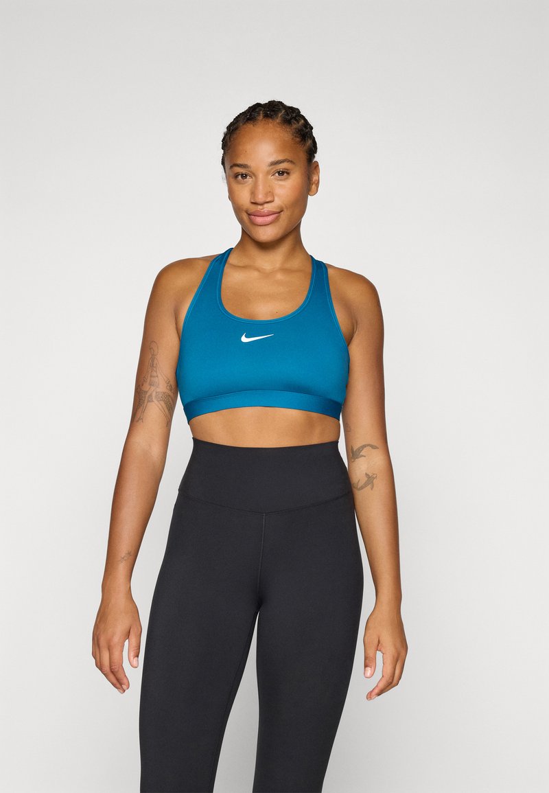 Una donna con i capelli intrecciati sorride, indossando un reggiseno sportivo verde acqua e leggings neri, mostrando una postura atletica sicura.