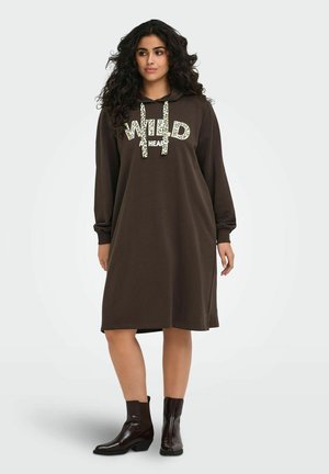 Žena s kučeravými vlasmi, ktorá má na sebe hnedé dlhé šaty s kapucňou s nápisom "WILD HEART" a tmavé členkové topánky, stojaca na jednoduchom pozadí.