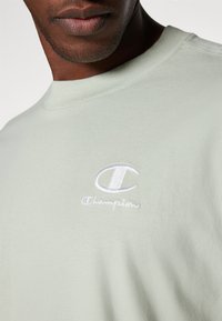 Lysegrøn bomulds t-shirt med rund hals, der har et hvidt, broderet Champion-logo på venstre bryst. Blød tekstur, enkelt design.