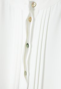 Witte blouse met knoopsluiting, voorzien van plooien en een gladde textuur. De knopen zijn iriserend in tinten crème en groen.