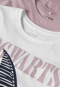Twee t-shirts opgestapeld, de ene wit met marineblauwe en roze graphics, de andere lichtroze. Beide hebben een ronde hals en zijn gemaakt van zachte katoenen stof.