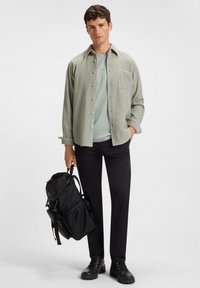 Man in een lichtgroene shirt over een groene t-shirt, zwarte broek, zwarte wandelschoenen, die een zwarte rugzak vasthoudt bij de handgreep.