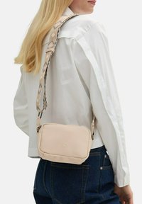 Borsa a tracolla beige con superficie testurizzata e tracolla decorata, indossata sulla spalla. Presenta una cerniera liscia e un dettaglio con logo.