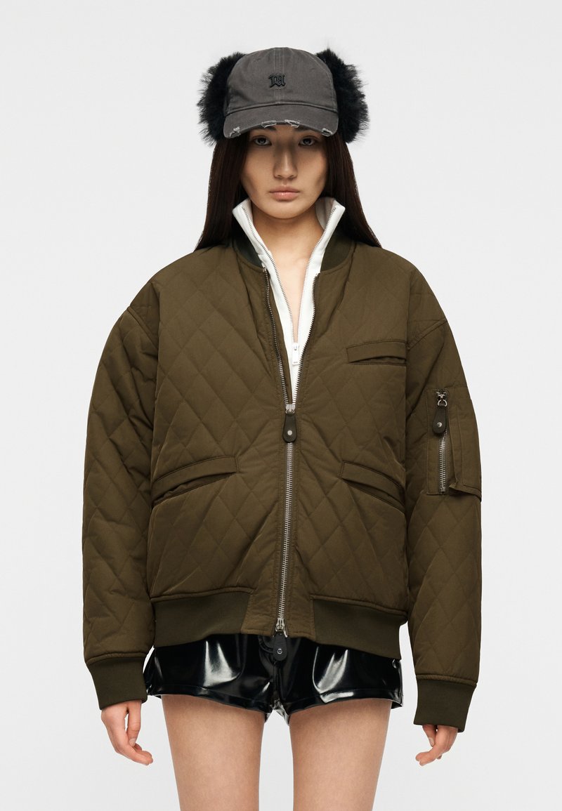 Γυναίκα που φοράει υπερμεγέθη χακί καπιτονέ μπο υφάν, μαύρα σορτς από ψεύτικο δέρμα και γκρι καπέλο με μαύρα γούνινα ακουστικά, στέκεται ουδέτερα μπροστά σε γκρι φόντο.