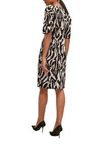 Femme portant une robe noire et blanche à motifs abstraits arrivant aux genoux avec des manches mi-longues et des talons hauts noirs, se tenant de dos.