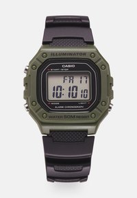 Montre numérique avec un boîtier en résine verte, un bracelet en caoutchouc noir et un afficheur LCD indiquant l'heure et la date. Les caractéristiques incluent une alarme et une résistance à l'eau.