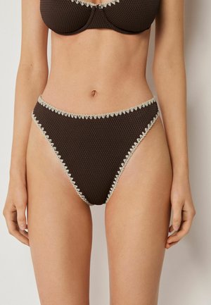 Bas de bikini texturé marron foncé taille haute avec bordure crème ondulée porté par une femme debout devant un fond neutre.
