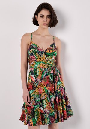 Ein Modell steht vor einem weißen Hintergrund und trägt ein buntes, blumenmusterndes Sommerkleid mit einem gerafften Saum und Spaghettiträgern.