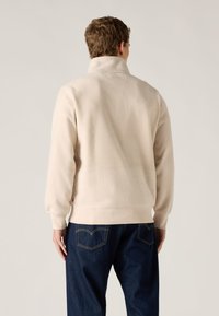 Φούτερ από μπεζ φλις με ψηλό γιακά και ριμπ μανσέτες, σε συνδυασμό με σκούρο τζιν παντελόνι με ορατές ραφές και τσέπες.