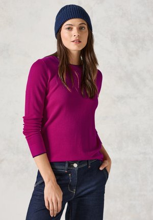 Fuchsia Langarmoberteil mit strukturiertem Stoff, rundem Halsausschnitt und taillierten Ärmeln, kombiniert mit dunkelblauen Jeans und einer marineblauen Strickmütze.