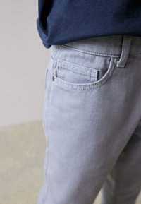 Vaqueros de mezclilla gris claro con diseño estándar de cinco bolsillos, con costuras visibles, detalles de remaches metálicos y textura suave.