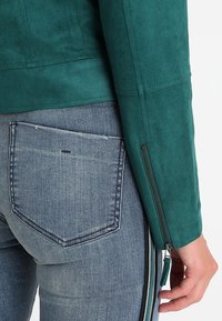 Zbliżenie osoby noszącej niebieskie dżinsy z tylną kieszenią oraz zieloną kurtkę z rękawem i detalem zamka błyskawicznego.