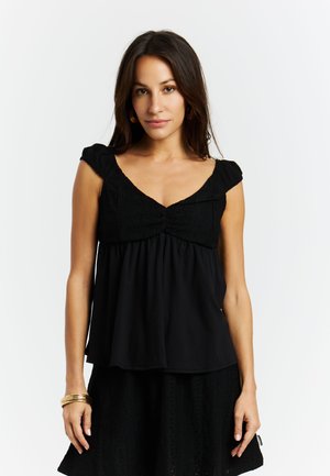 Jeune femme portant un haut sans manches noir à col en V et une jupe, debout devant un fond blanc uni, avec des boucles d'oreilles créoles dorées et des bracelets.