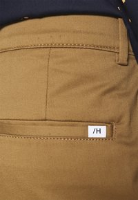Pantaloni in cotone marrone con una texture liscia e taglio dritto. Presentano una tasca posteriore con una piccola etichetta bianca con la scritta "/H".