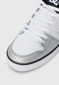 Zapatilla blanca con puntera texturizada en metal plateado, cordones blancos y suela con ribete negro sobre una superficie clara.