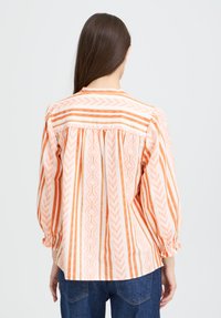 Femme avec de longs cheveux bruns portant un blouse ample à manches longues avec des rayures verticales orange et blanches, associée à un jean bleu.