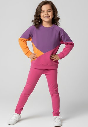 Uśmiechnięta dziewczyna z falującymi włosami stoi z rękami na biodrach, ubrana w bluzę z blokami kolorów fioletowego, pomarańczowego i różowego, różowe spodnie i białe trampki.