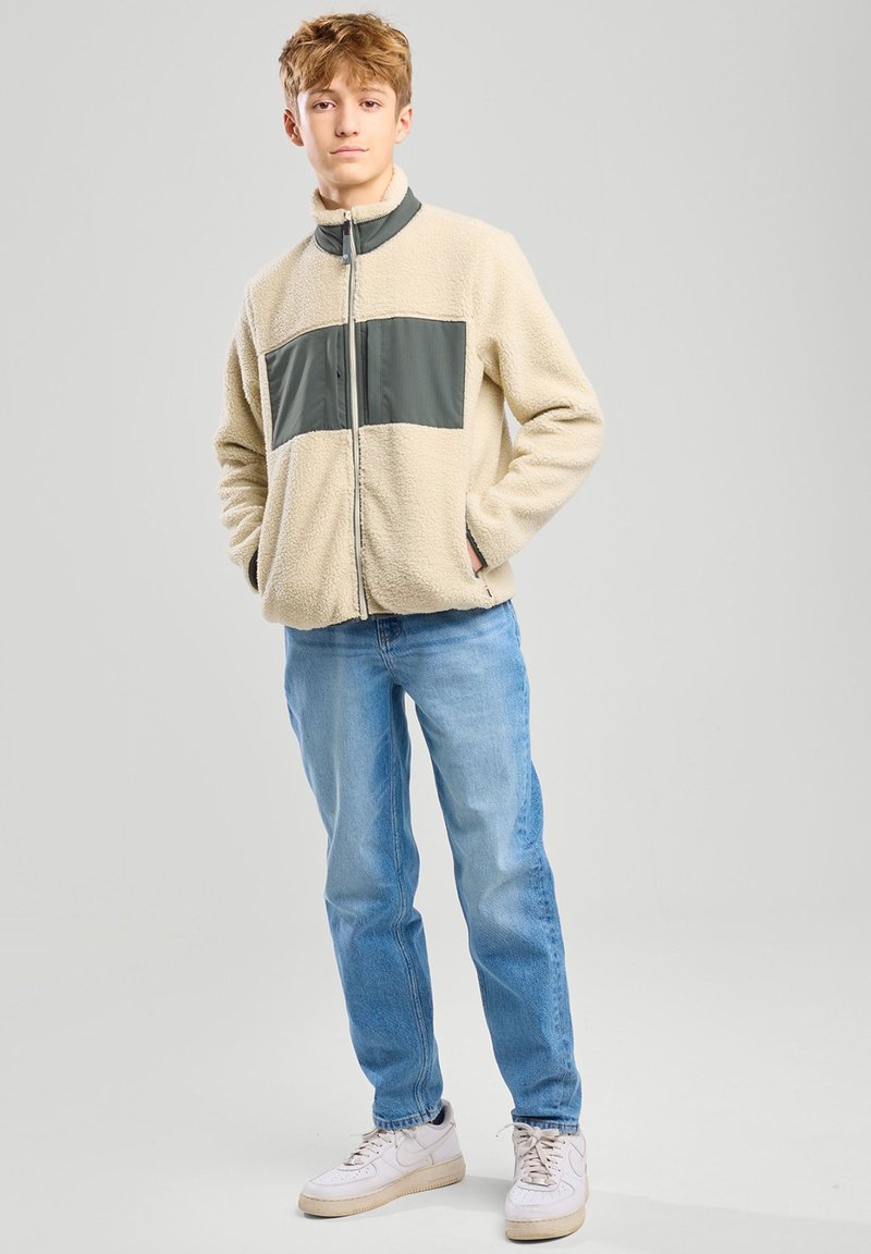 Giacca in pile beige con pannello grigio, zip centrale, tasche laterali, abbinata a jeans azzurri chiari e sneaker bianche su uno sfondo neutro.