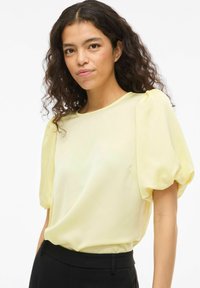 Blouse jaune à manches courtes bouffantes, col rond et texture lisse, associée à un pantalon noir. Design simple sans motifs visibles.