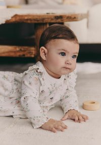 Bebé con un pelele de estampado floral claro y hombreras con volantes, gateando sobre una superficie texturizada, junto a un juguete de anillo de madera.