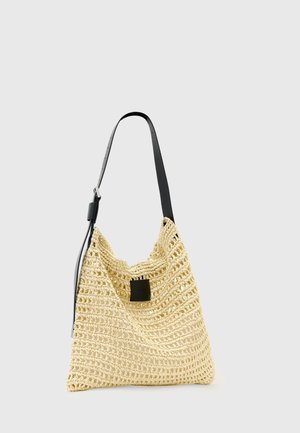 Sac fourre-tout beige tissé avec une bandoulière noire réglable et un petit patch rectangulaire noir sur le devant.