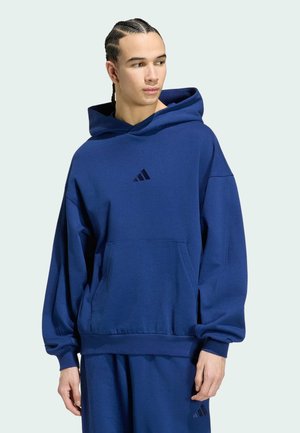 Młody mężczyzna z plecionymi włosami, ubrany w luźną niebieską bluzę Adidas z kapturem i pasujące spodnie dresowe, patrzy w lewo na jednolitym tle.