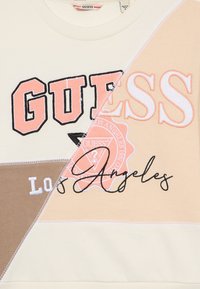 Sweatshirt crème et beige avec une grande inscription "GUESS", des accents brodés et un graphique "Los Angeles" en cursive sur un tissu doux.