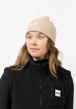Beige gebreide beanie met een geribbelde structuur, voorzien van een vierkant label. Gecombineerd met een zwarte fleece vest met een ritszak.