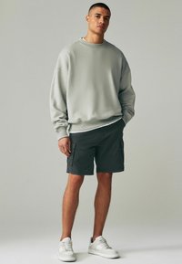 Hellegrauer, übergroßer Pullover, dunkle Cargo-Shorts und weiße Sneakers. Texturierter Pullover mit gerippten Bündchen; die Shorts haben Seitentaschen.