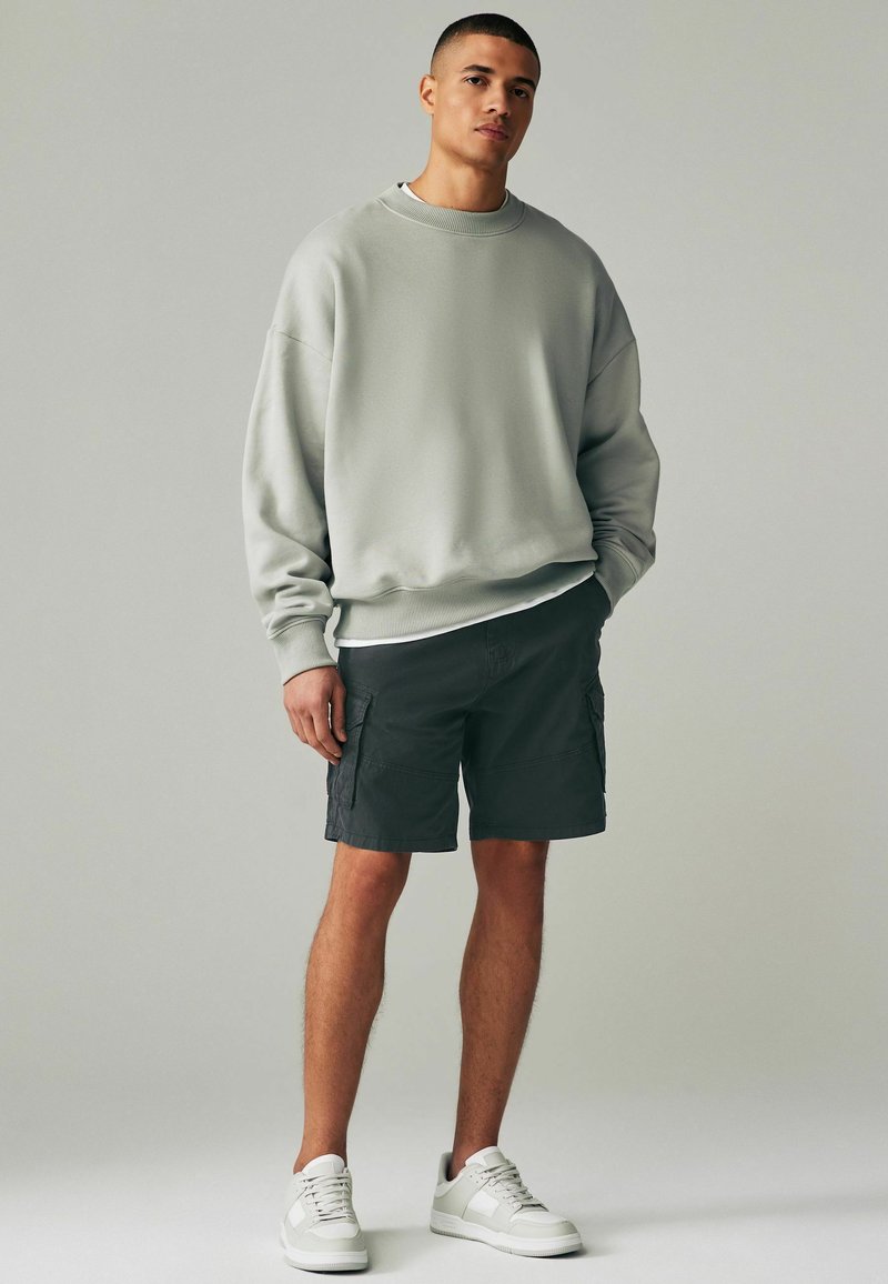Hanorac oversized gri deschis, pantaloni scurți cargo negri și adidași albi. Hanoracul are o textură cu manșete ribbed; pantaloni scurți cu buzunare laterale.