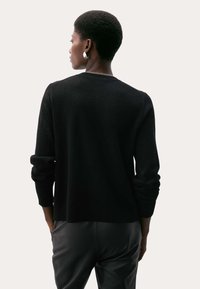 Černý svetr z vlněné směsi s kulatým výstřihem, žebrovanými manžetami a detaily na zadní straně. Kombinován s černými přiléhavými kalhotami. Hladká textura.