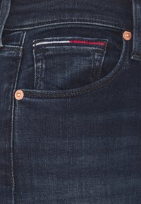 Jeans de mezclilla azul oscuro con un pequeño bolsillo frontal que presenta costuras en rojo, blanco y azul, y remaches de cobre en las costuras.