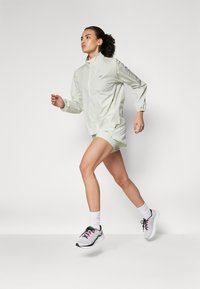 Casaco de corrida verde claro com punhos elásticos, combinado com calções verdes claros. Ténis brancos e pretos com detalhes em rosa. Posição de corrida.