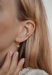 Orecchini ad anello in oro con design floreale, decorati con cristalli trasparenti. Indossati su un orecchio forato, che completano capelli lunghi e lisci.