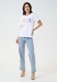 Un t-shirt blanc avec un motif pixelisé coloré en rouges et bleus, associé à un jean large bleu clair et des bottines beige.